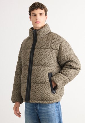 Quilted jacket in olive green fabric with a raised pattern, high collar, and black zipper details. Features side pockets and a relaxed fit.