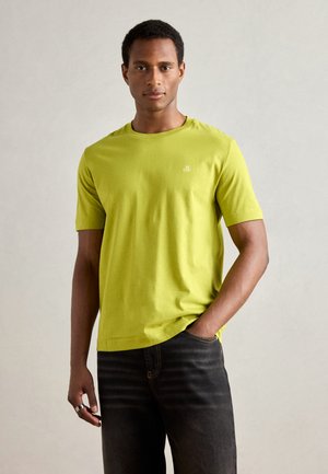 Camiseta de algodón verde lima con mangas cortas y un cuello redondo. Presenta un pequeño logotipo blanco en el lado izquierdo del pecho. Combinada con jeans oscuros.