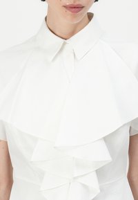 White blouse featuring a button-down collar and layered ruffle design at the front. Made of a smooth fabric with subtle sheen.