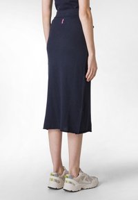 Fustă bleumarin, până la genunchi, cu un elastic texturat la talie, textură netedă, slit lateral și detalii cu etichetă roz. Asortată cu pantofi sport gri deschis.