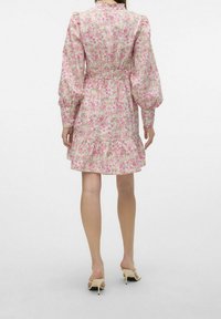 Kvinna klädd i en långärmad rosa blommig klänning med samlad midja och volangkant, tillsammans med beige öppna högklackade sandaler, stående.