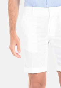 Pantaloni corti bianchi in lino con taglio dritto, caratterizzati da un orlo risvoltato e decorazioni minime, abbinati a una camicia azzurra chiara sullo sfondo.