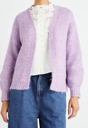 Kvinde iført en tekstureret lavendel cardigan med åben front over en hvid bluse med blondeknapper og blå høj-taljede jeans.