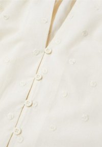 Vaalea kermainen paita, jossa on teksturoitu kukkakirjailu, nappikiinnitys edessä ja useita pieniä nappeja. Pehmeää kangasta, jossa sileä viimeistely.