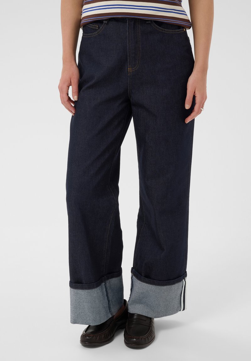 Personne portant un jean large foncé à grands revers, des mocassins foncés et un haut rayé marron et blanc, debout devant un fond uni.