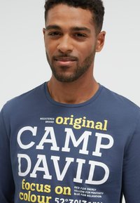 Mann mit kurzen lockigen Haaren und Bart, trägt ein marineblaues Hemd mit dem weißen und gelben Schriftzug "original CAMP DAVID focus on colour".