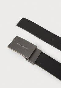Cinturón negro con acabado mate, que presenta una hebilla metálica rectangular grabada con "Armani Exchange". Diseño simple, longitud ajustable.