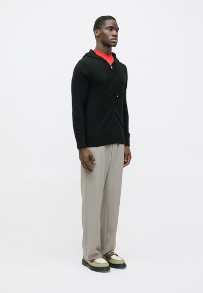 Homem em hoodie preto com fecho, camisa vermelha, calças largas beje e sapatos mocassins bicolores, posicionado contra um fundo branco liso.