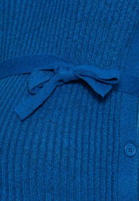 Cardigan bleu tricoté présentant une texture côtelée, une ceinture nouée à la taille et un détail de bouton bleu sur le côté.