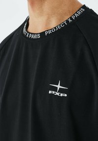 Camiseta negra de manga corta con cuello redondo, que presenta un logo blanco contrastante "PXP" y una marca a lo largo del cuello. Tejido de algodón suave.
