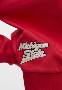 Sweat-shirt rouge avec texte brodé "Michigan State" en blanc et noir, affichant un design incurvé distinct en bas.
