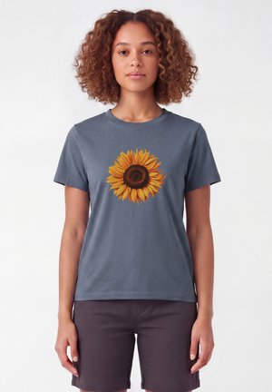Femme aux cheveux bouclés portant un t-shirt bleu-gris avec un grand motif de tournesol, associée à un short marron foncé, debout devant un fond blanc.