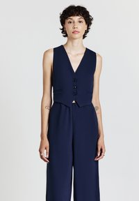 Combinaison sans manches bleu marine avec un décolleté en V, cinq boutons et deux poches avant ; fabriquée à partir d'un tissu lisse avec une coupe ajustée.