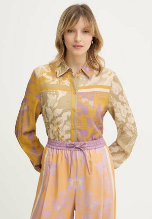 Donna con capelli biondi fino alle spalle che indossa una camicia a fantasia a bottoni nei colori senape, beige e lilla, abbinata a pantaloni a vita alta dello stesso motivo.