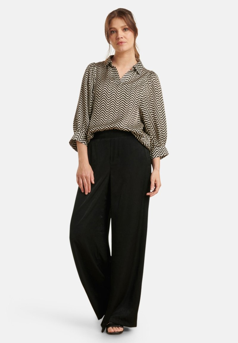 Zwart-witte blouse met chevronpatroon en kraag, gecombineerd met hoge zwarte wijde broek en platte sandalen.