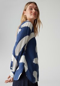 Marineblaue Bluse mit weißem abstraktem Pinselstrichmuster, langen Ärmeln, rundem Saum und geknöpften Manschetten. Glatter Stoff mit leichtem Glanz.