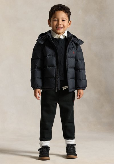 Niño pequeño sonriente, vestido con una chaqueta acolchada negra, suéter negro, camisa blanca, pantalones negros, calcetines blancos y zapatillas negras, de pie sobre un fondo neutro.