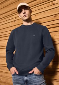 Navy blue sweatshirt with a round neckline, long sleeves, and a small logo on the chest; worn with light blue jeans and a beige cap.