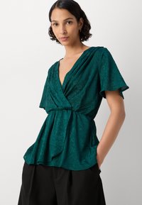 Blusa teal con scollatura a V profonda, maniche corte e vita regolabile con stringhe, caratterizzata da un motivo testurizzato e un tessuto morbido e fluente.