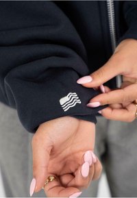 Námornícky svetri s rukávom, ktorý je ozdobený bielym vyšitým vzorom vlny. Hladký bavlnený materiál s priliehajúcim manžetovým finišom. Ruka ukazuje detailne upravené nechty.