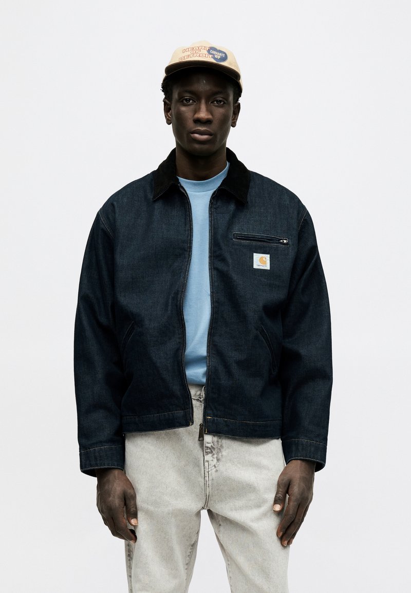 Joven con gorra beige, chaqueta de mezclilla oscura con bolsillo en el pecho con cremallera, camisa azul claro y vaqueros gris claro sobre un fondo blanco.