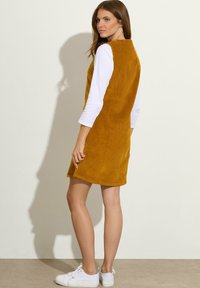Vestido sin mangas de pana mostaza con cuello alto, que presenta bolsillos laterales; superpuesto a una camiseta blanca de manga larga. Zapatillas blancas abajo.