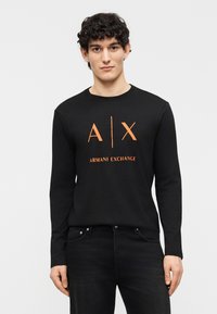 Νεαρός άνδρας με σγουρά μαλλιά που φοράει μαύρο μακρυμάνικο πουκάμισο Armani Exchange με πορτοκαλί λογότυπο, σε συνδυασμό με μαύρο τζιν, στέκεται μπροστά σε απλό φόντο.