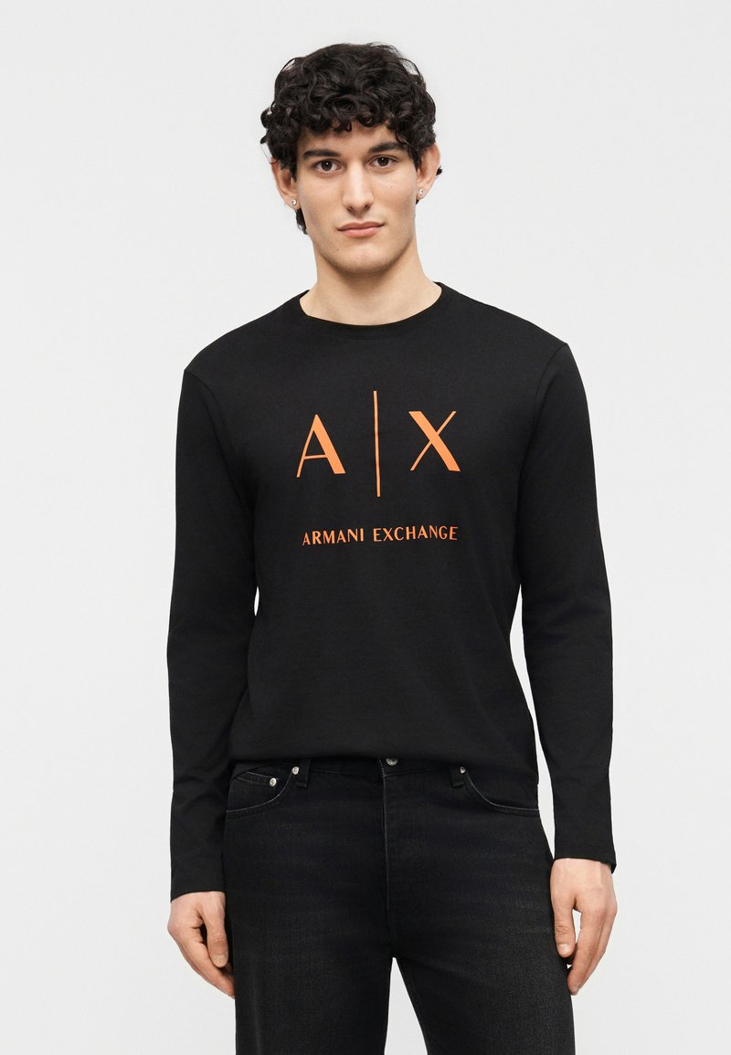 Νεαρός άνδρας με σγουρά μαλλιά που φοράει μαύρο μακρυμάνικο πουκάμισο Armani Exchange με πορτοκαλί λογότυπο, σε συνδυασμό με μαύρο τζιν, στέκεται μπροστά σε απλό φόντο.