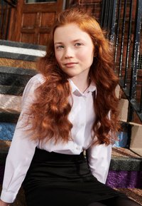 Camicia bianca a maniche lunghe e gonna nera, seduta su gradini colorati. Capelli rossi lunghi e ondulati, pelle chiara e espressione neutra.