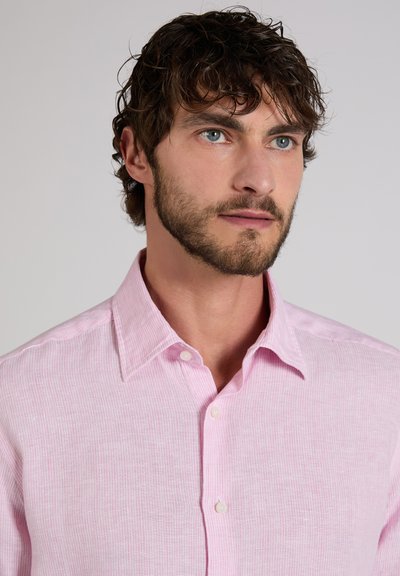 Uomo con capelli ricci castani e barba, che indossa una camicia rosa chiaro abbottonata, che guarda leggermente di lato su uno sfondo semplice.