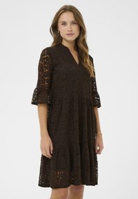 Ruda nėriniuota suknelė su V formos iškirpte, varpų formos rankovėmis ir sluoksnių sijonu. Yra gėlių raštas ir pusiau permatomos tekstūros.