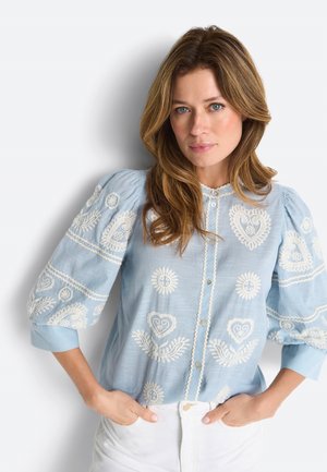 Frau mit welligem Haar, die eine hellblaue Bluse mit weißen, gestickten Herzen und Blumen trägt, die Hände in den Taschen einer weißen Hose vergraben.