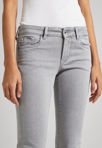 Mujer vistiendo jeans ajustados de color gris claro y una camiseta de tirantes blanca con textura, de pie con las manos relajadas a los lados frente a un fondo neutro.