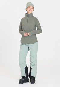Fleece pullover i mørkegrøn, design med halv lynlås. Lysegrønne snebukser med sorte accenter, parret med sorte skistøvler og en lysegrå strikket hue.