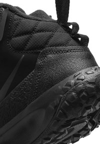 Primer plano de la parte trasera de una zapatilla deportiva negra que muestra el tejido cosido, la suela de goma texturizada con un diseño rugoso y una lengüeta en la parte posterior.