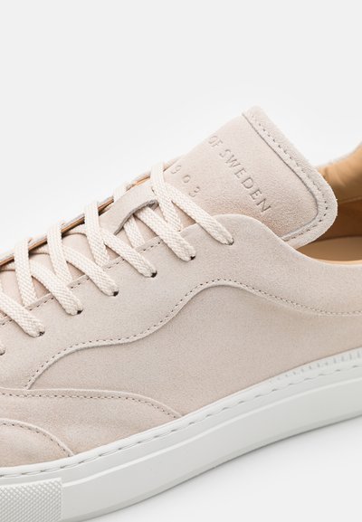 Zapatillas de ante beige con una parte superior texturizada, cordones planos y una suela de goma blanca; presenta un logotipo en relieve en la lengüeta.