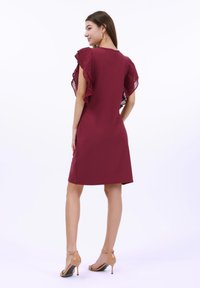 Burgundy klänning med volangärmar, figursydd form och knälång design. Slätt tyg med genomskinliga inslag. Buren med beige högklackade sandaler.