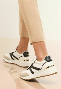 Sneaker bianchi con dettagli neri testurizzati e hardware dorato. Suola spessa, design a pannelli lisci e a contrasto. Indossati con pantaloni beige tagliati.