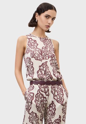 Kvinde iført ærmeløs top og bukser med store abstrakte vinrøde løvmønstre på hvid, hænder i lommerne, flettet vinrødt bælte.