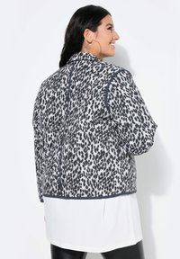 Vatteret jakke i grå med sort leopardprint, med en rund krave, elastiske mansjetter og marineblå trim. Bæret over en hvid skjorte.