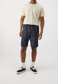 Conjunto masculino que incluye una camiseta de cuello redondo de color beige claro y pantalones cortos azul marino con un patrón de cuadrícula azul, combinados con zapatillas negras y calcetines blancos.