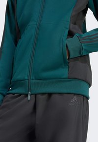 Teal og sort lynlåsjakke med tekstureret stof, der har en kontrastsort panel, elastiske manchetter og adidas-logoet på bukserne.