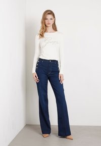 Camicetta bianca a maniche lunghe con "Guess" in strass, abbinata a jeans a gamba larga blu scuro con dettagli in bottone sulle tasche.