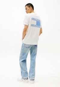 Hvid bomulds T-shirt med blå mønstret bagprint, parret med lyseblå denimjeans. Outfittet har en afslappet pasform og minimal detaljer.