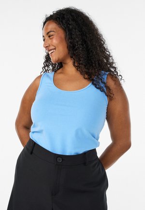 Lächelnde Frau mit lockigem Haar, die ein hellblaues ärmelloses Oberteil und schwarze Hose vor weißem Hintergrund trägt.