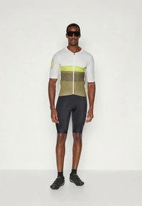 Jersey de ciclismo de manga corta en blanco con rayas horizontales verdes y olivas, combinado con pantalones cortos negros, con una cremallera frontal y textura de malla.