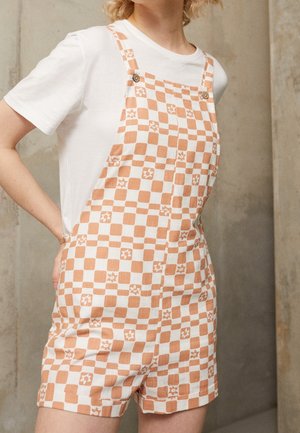 Mono corto a cuadros en tonos durazno y blanco, con estampados florales, tirantes ajustables y un corte relajado, usado sobre una camiseta blanca.