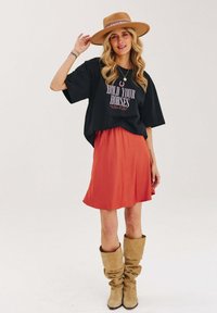 Čierne oversized tričko s grafikou "HOLD YOUR HORSES", spárované s oranžovou sukňou. Hnedé voľné čižmy a široký klobúk dopĺňajú vzhľad.