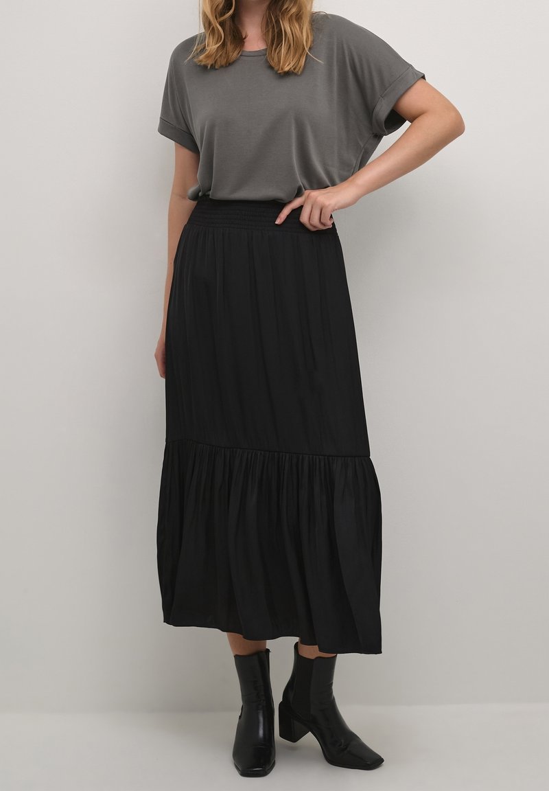 Šedý top s krátkým rukávem v kombinaci s dlouhou černou sukní s volány. Sukně má smockingový pas a splývavou látku. Černé kotníkové boty celý outfit doplňují.