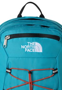 Teal-nahkainen reppu, jossa musta vetoketju ja yksityiskohdat. Varustettu valkoisella logolla ja joustavalla nyörillä. Kompakti ja jäsennelty muotoilu.